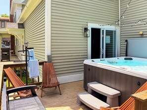 A hot tub on a deck with wooden chairs and a table, a door to a covered area, and string lights. - Downtown - Hot Tub, Speakeasy, Grill, Game Room, Fireplace (Ludlow)