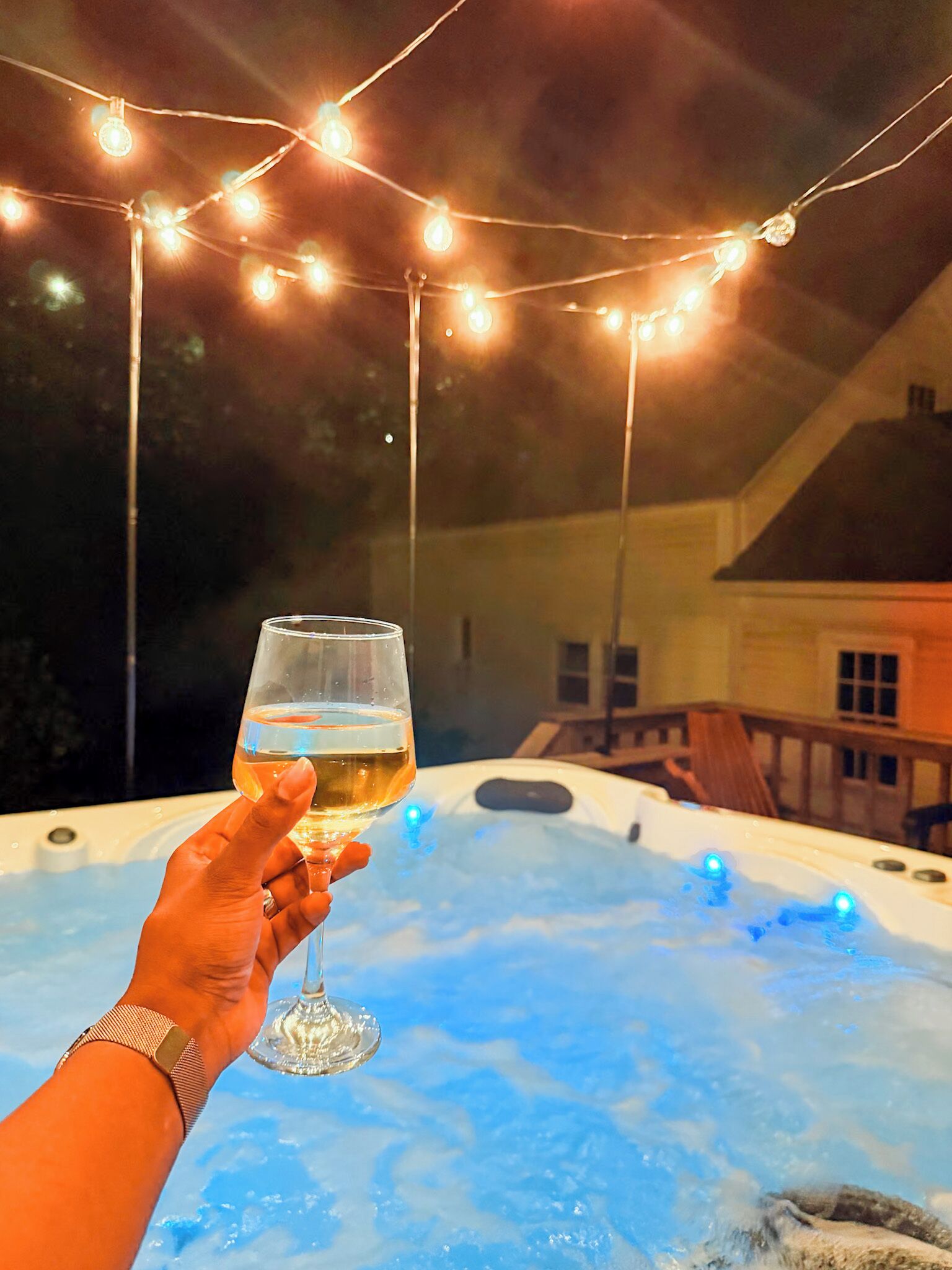 A person holding a glass of white wine with a hot tub in the background, string lights overhead, and a house visible in the distance.