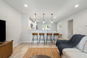 A modern living room with a sofa, a TV on a wooden cabinet, a kitchen area with white cabinets, a bar stool, and a ceiling with recessed lighting. - New 4 Bed 2 and a Half Bath Near ND Campus (South Bend)