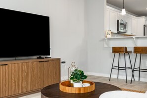 Ein modernes Wohnzimmer mit Flachbildfernseher, einer Holz-Unterhaltungseinrichtung, einem runden Couchtisch mit Pflanze und einer Küchenzeile mit weißen Schränken und Barhocker.