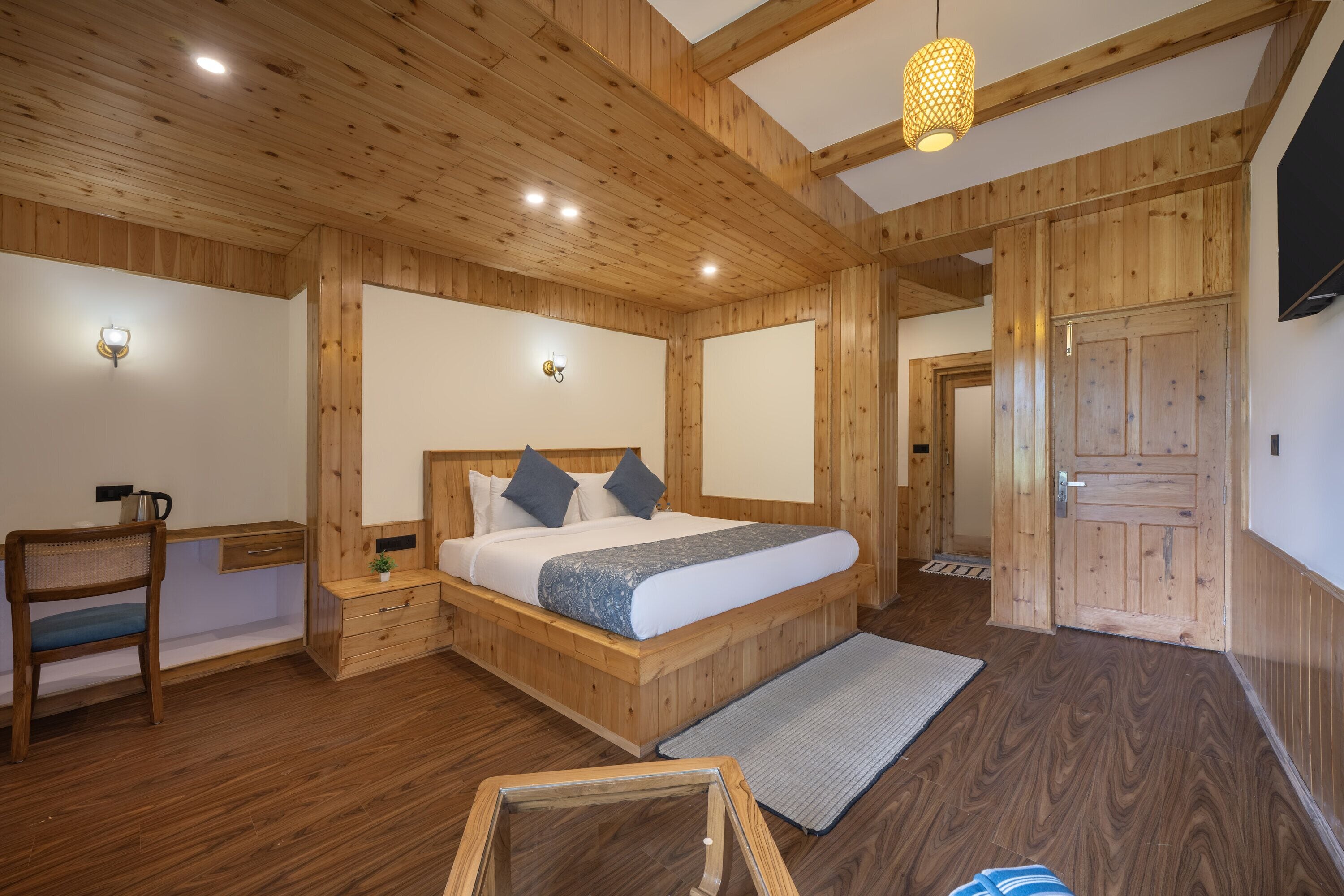 A wooden room with a bed, bedside tables, a chair, a desk, and a door.