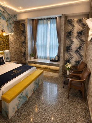 Una habitación de hotel con una cama, un área de descanso con sillas, una mesita con un florero y una ventana con cortinas.