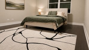 A bedroom with a bed, a nightstand, a lamp, a window with blinds, and a painting on the wall.