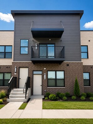 A modern two-story building with a flat roof, a balcony on the second floor, and a front entrance with a door and a small set of stairs. - Furnished 1BR - Huntley Crossing Luxury Apartment (Huntley)