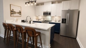 A modern kitchen with a central island, bar stools, a stainless steel refrigerator, and a hanging light fixture.