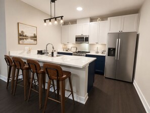 A modern kitchen with a central island, bar stools, a stainless steel refrigerator, and a hanging light fixture. - Furnished 1BR - Huntley Crossing Luxury Apartment (Huntley)