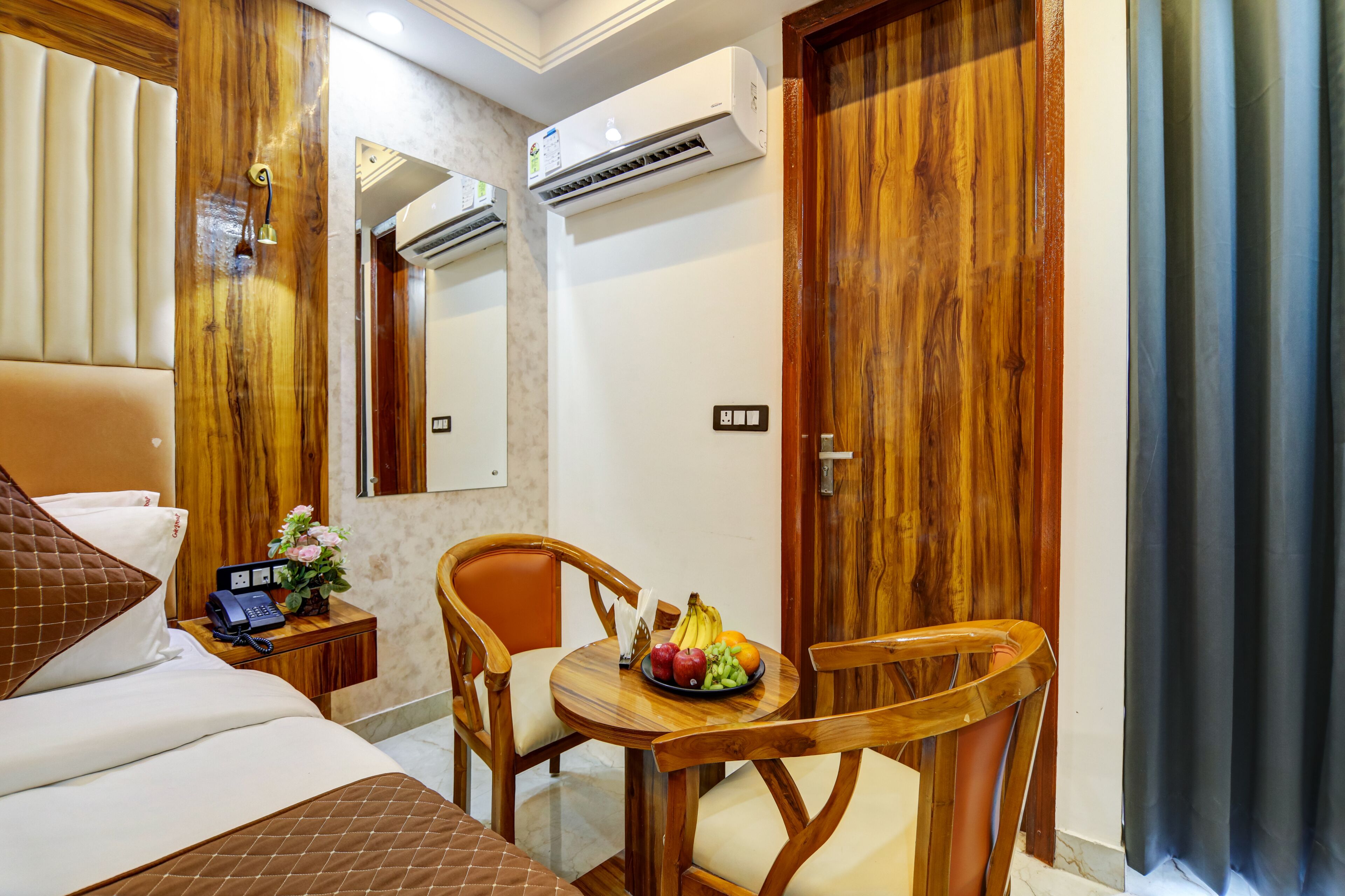 A hotel room with a bed, a desk with a telephone and a vase of flowers, a chair, a round table with fruit, and wooden doors.