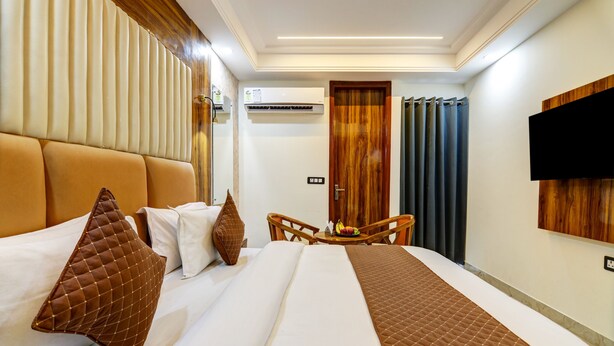 A hotel room with a large bed, a wooden headboard, a flat-screen TV, a small table with fruit, and a door with a wooden panel. - Hotel Signature Suites (New Delhi)