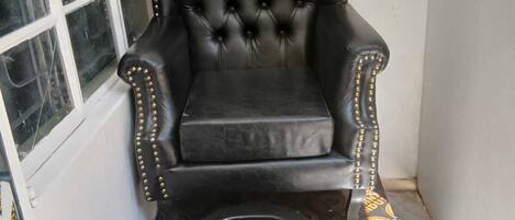Un sillón de cuero negro con tachuelas doradas, un sillón de masaje negro con detalles dorados y un ventanal con marcos blancos.