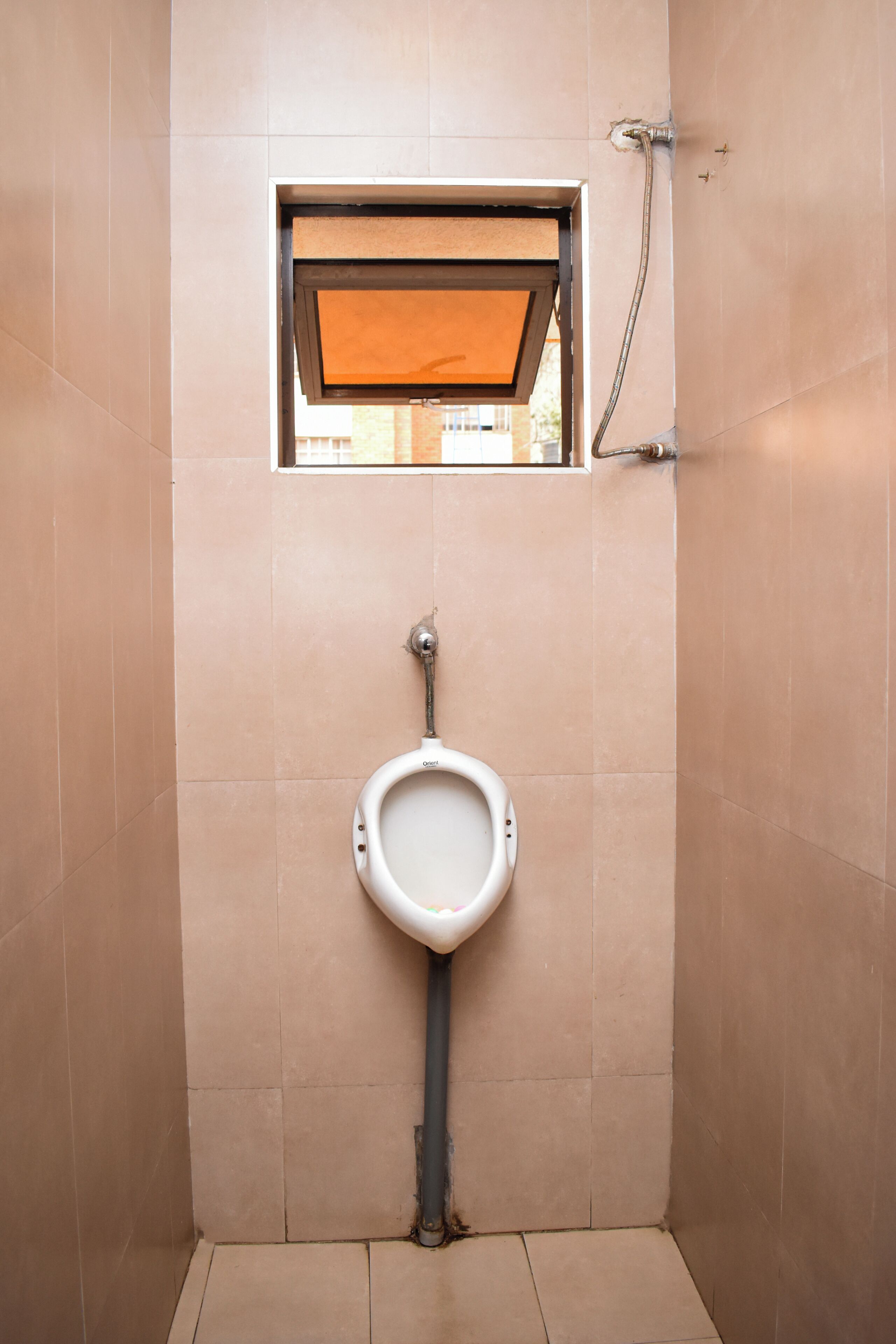 A urinal with a window above it, a showerhead to the right, and tiled walls.
