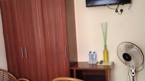 A room with a TV mounted on the wall, a wardrobe, two wicker chairs, a small table with a bottle and a remote, and a standing fan. - Jp Resort & Spa (Nairobi)