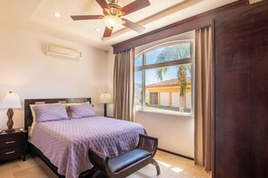 Een slaapkamer met een bed, twee nachtkastjes, een bureau, een stoel, een plafondventilator, een raam met gordijnen en een airconditioning.