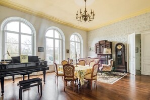 A spacious room with a grand piano, a dining table with chairs, and a large arched window.