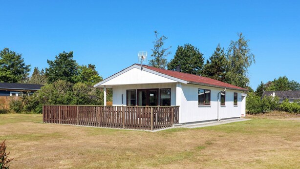 A single-story building with a white exterior and a red roof, surrounded by a wooden fence and a grassy yard. - 5 Person Holiday Home in Hadsund (Hadsund)