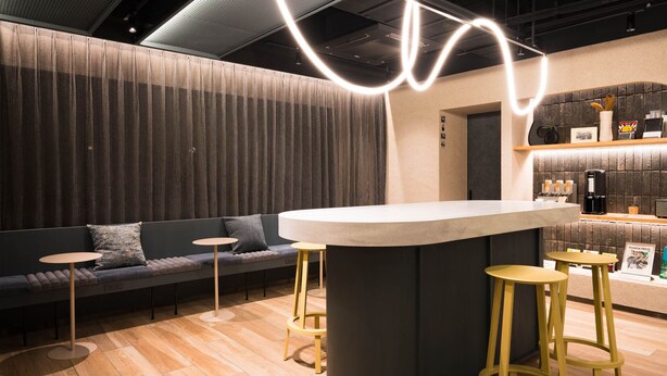 A modern bar area with a round bar counter, two stools, a sofa with cushions, and a wall with a textured pattern. - R Hotel Nagoya Station (Nagoya)