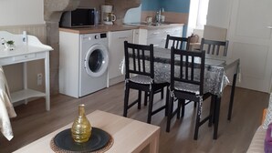 Een compacte keuken met een wasmachine, een eethoek met stoelen en een tafel, en een klein decoratief vaasje op de tafel.