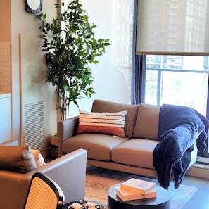 Una sala de estar con sofá, una mesa de centro, una planta y una ventana con vistas a edificios.