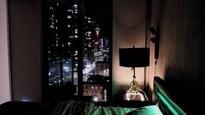 Un dormitorio con vistas a un paisaje urbano nocturno, una mesita de noche con lámpara y una cama con ropa de cama verde.
