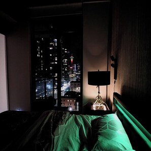 Quarto com vista para a cidade à noite, mesa de cabeceira com abajur e cama com roupa de cama verde.