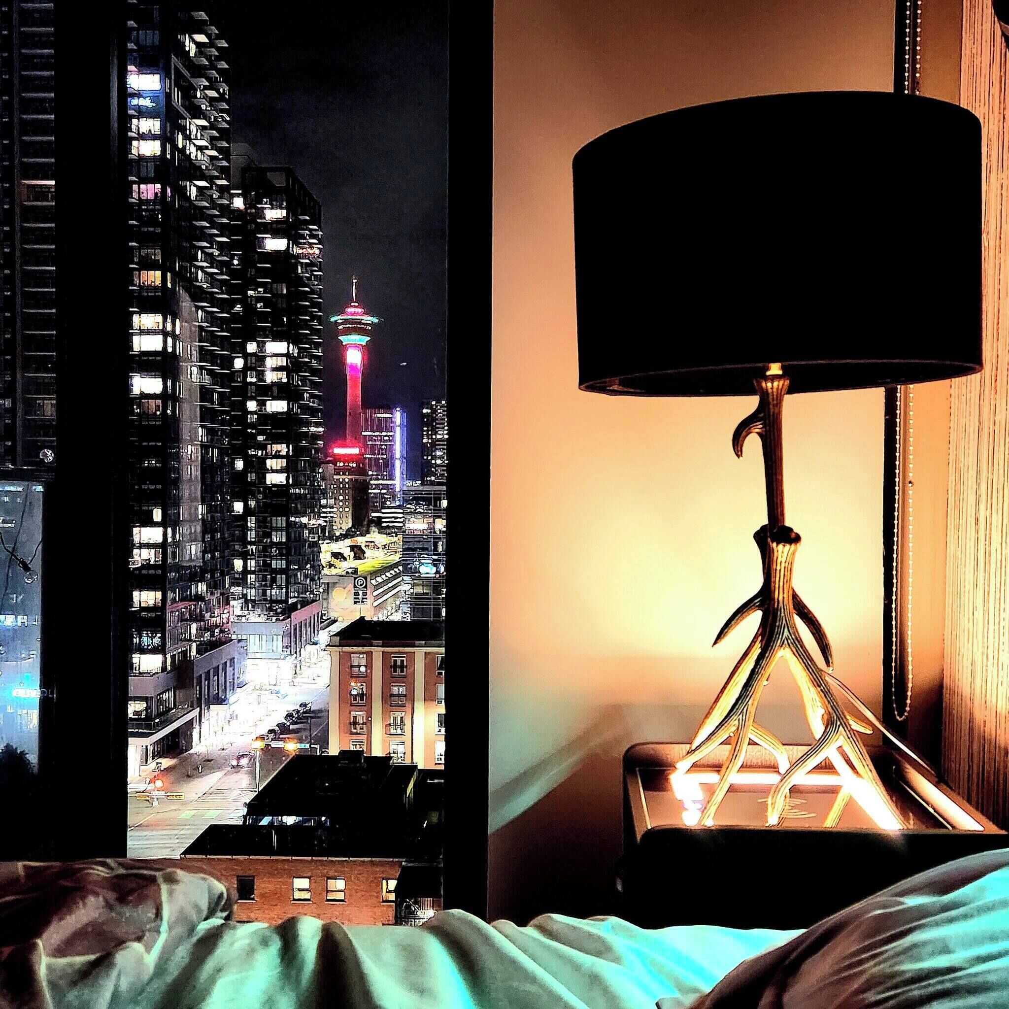 A bed with a view of a cityscape at night, including a tower with colorful lights.