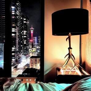 Una cama con vista a un paisaje urbano nocturno, incluyendo una torre con luces de colores.