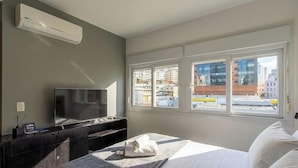 A hotel room with a bed, a flat-screen TV, a desk with a computer, and a window with a city view. - Studio in Condo w/ Pool in POA Pue0308 (Porto Alegre)