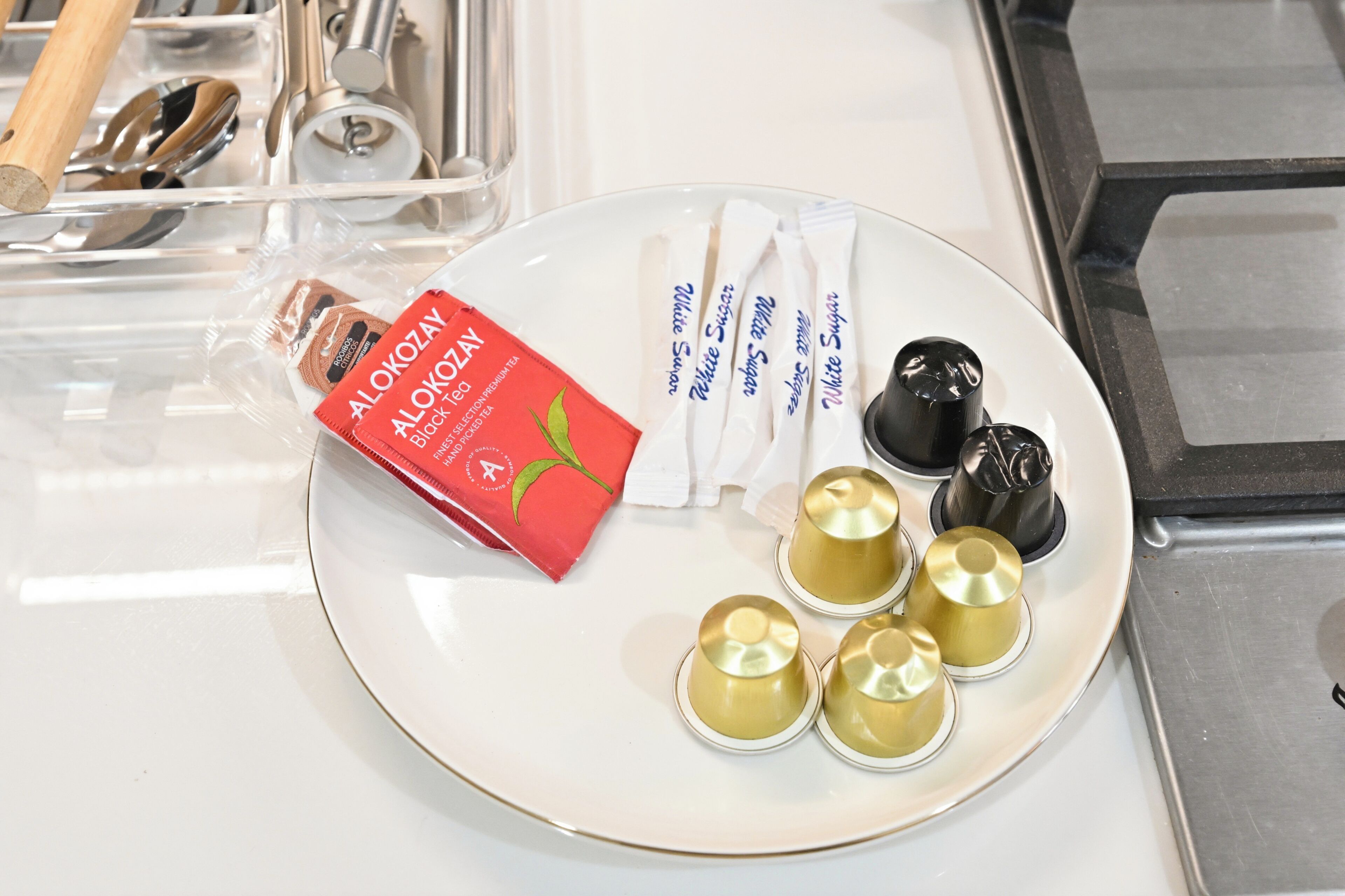 A plate with a red box labeled 'AloKozay Black Tea,' white packets, and four golden cups on a white surface.