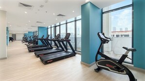 Ein Fitnessstudio mit Laufbändern, die an einer Wand mit großen Fenstern entlanggestellt sind, durch die Tageslicht und die Stadtblick möglich sind.