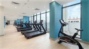 A gym with treadmills aligned against a wall with large windows, allowing natural light and views of the city. - Bloomfields Studio in Azizi Riviera 34 (Dubai)