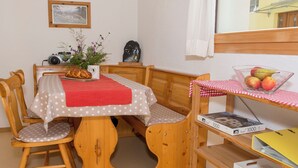 Et hyggeligt rum med et træbord, stole, en lille reol med en skål frugt, et ur og et billede i ramme på væggen.