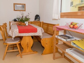 A cozy room with a wooden dining table, chairs, a bench, a small shelf with fruits, a clock, and a framed picture on the wall.