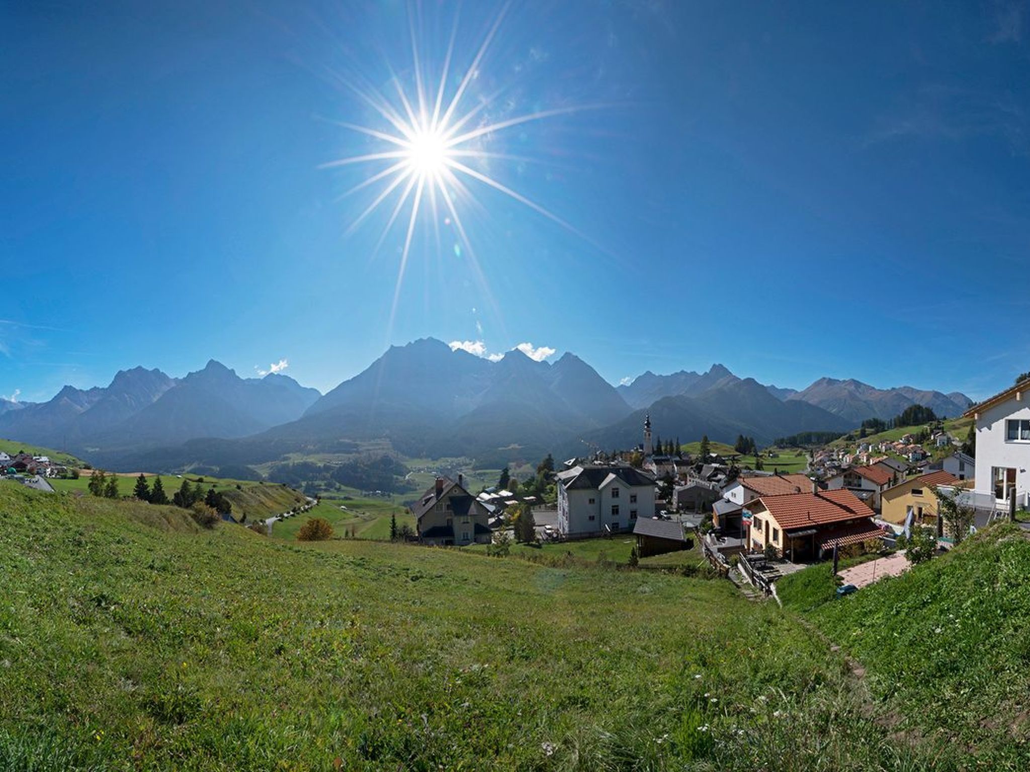 Ein Dorf mit Häusern und einer Kirche, umgeben von Bergen und wolkenlosem Himmel.