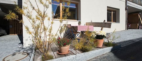 Rakennuksessa on päällystetty sisäänkäynti, ruukuissa olevia kasveja, ruutukuosinen pöytäliina ja puinen penkki.