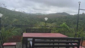 A table and chairs set outdoors with a view of a hilly landscape. - Vagadrops (Peermade)
