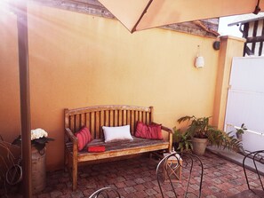 A wooden bench with cushions, a potted plant, and a metal chair in a courtyard with a tiled floor. - La Parenthèse d'Honfleur ~ 4 Bedrooms & Private Courtyard (La Rivière-Saint-Sauveur)