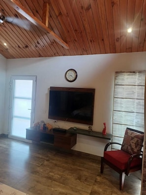 A room with a wooden ceiling, a flat-screen TV mounted on the wall, a door with a glass panel, and a red chair.