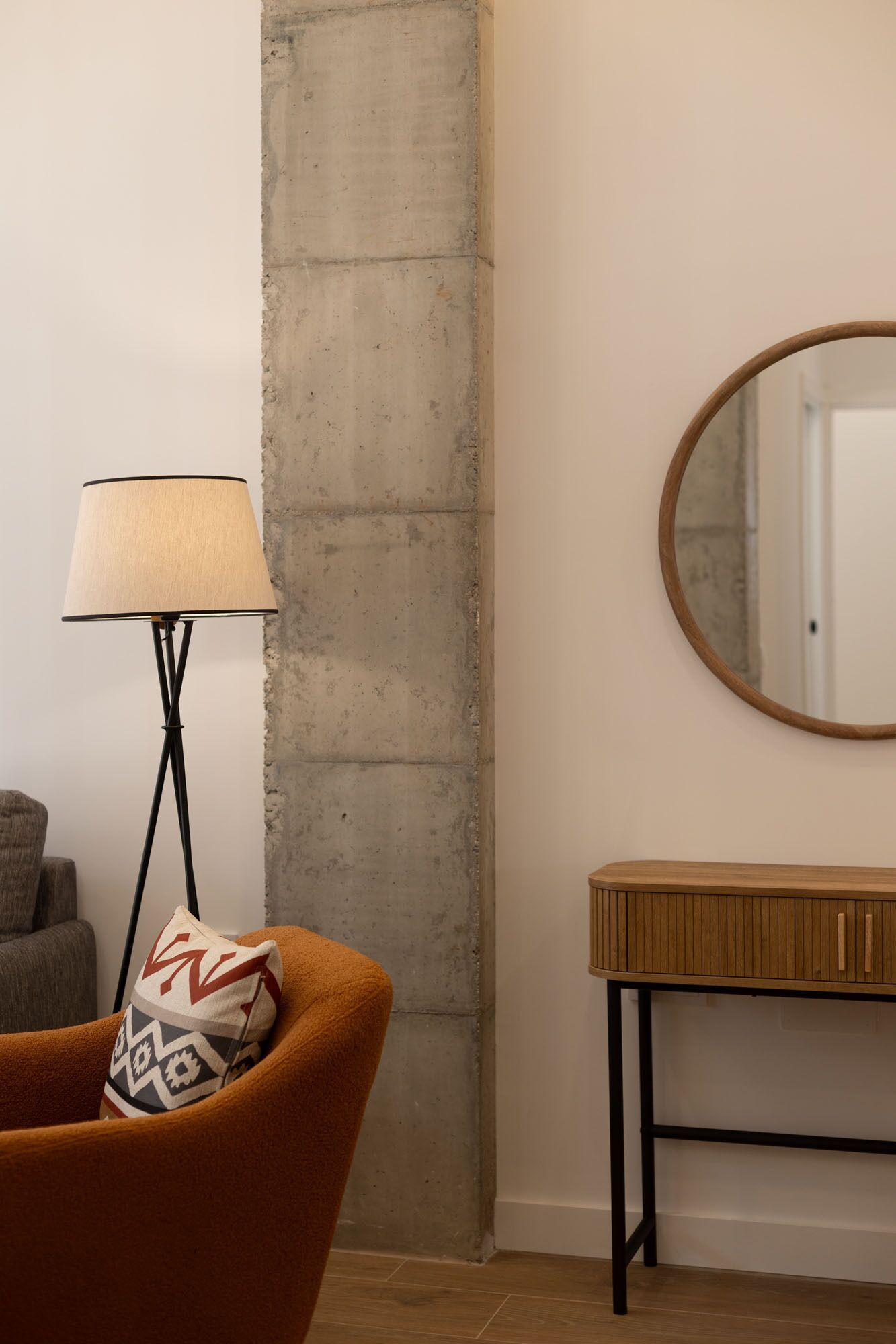A room with a concrete accent wall, a floor lamp, a round mirror, a wooden console table, and a patterned throw pillow.