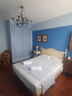A bedroom with a bed, a wooden nightstand, a chair, a wardrobe, and a chandelier.