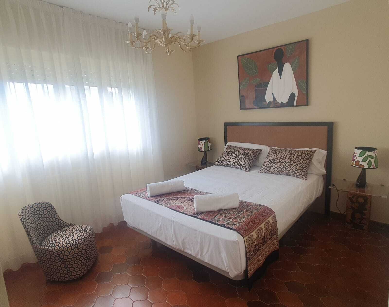 Un dormitorio con cama, una silla, una lámpara, un cuadro y una araña.