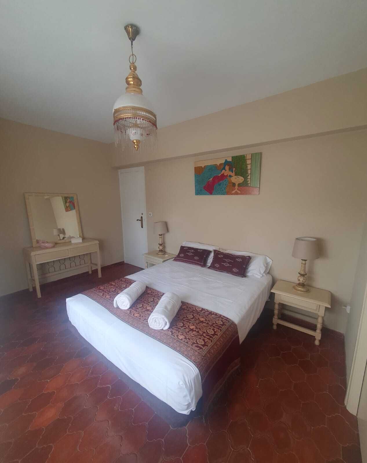A bedroom with a large bed, two bedside tables, a chandelier, a painting, and a mirror.