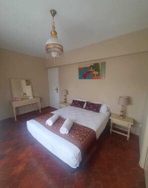 A bedroom with a large bed, two bedside tables, a chandelier, a painting, and a mirror.