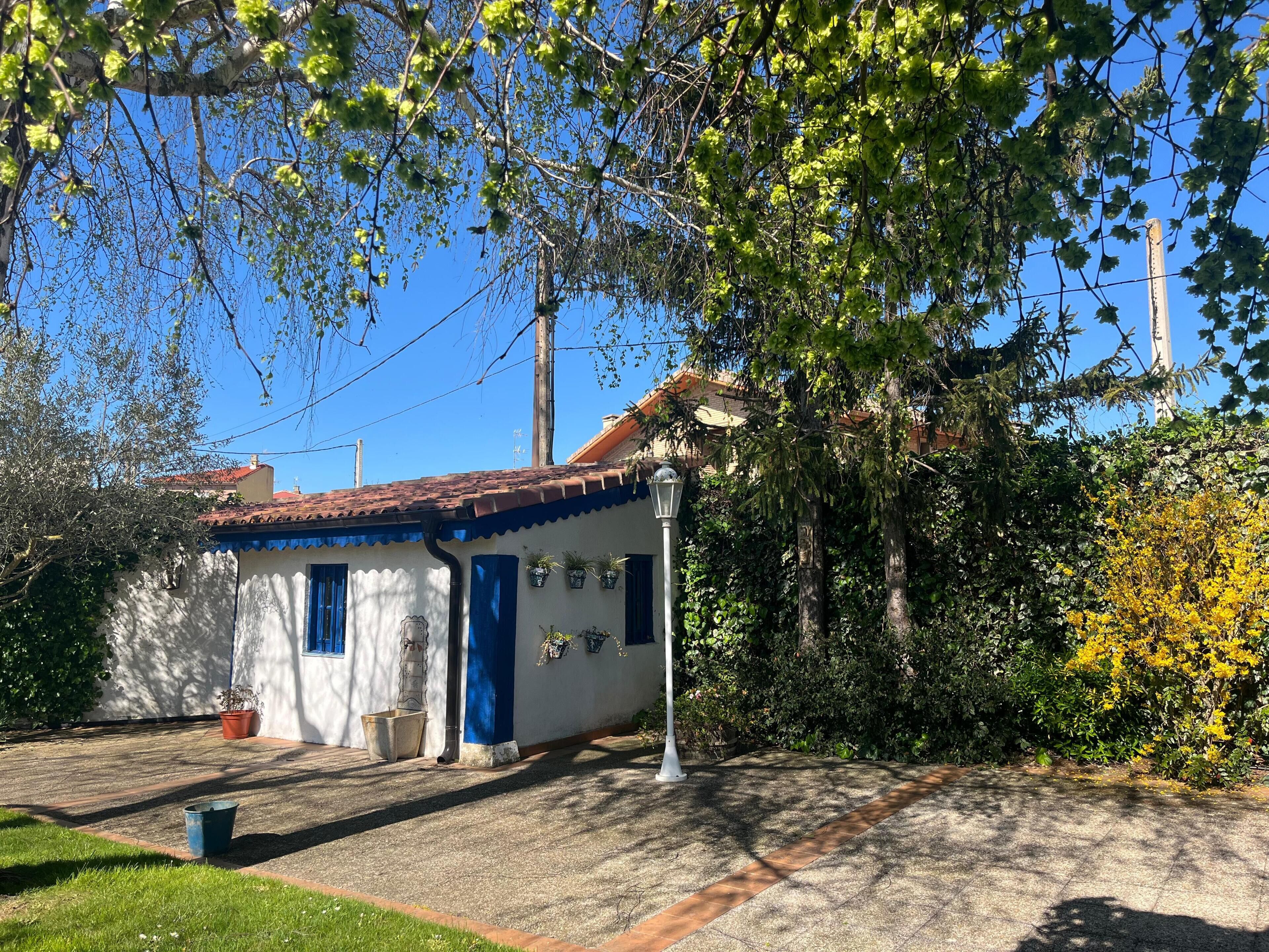 Un edificio blanco con detalles azules y techo de tejas, rodeado de vegetación y un cielo despejado.