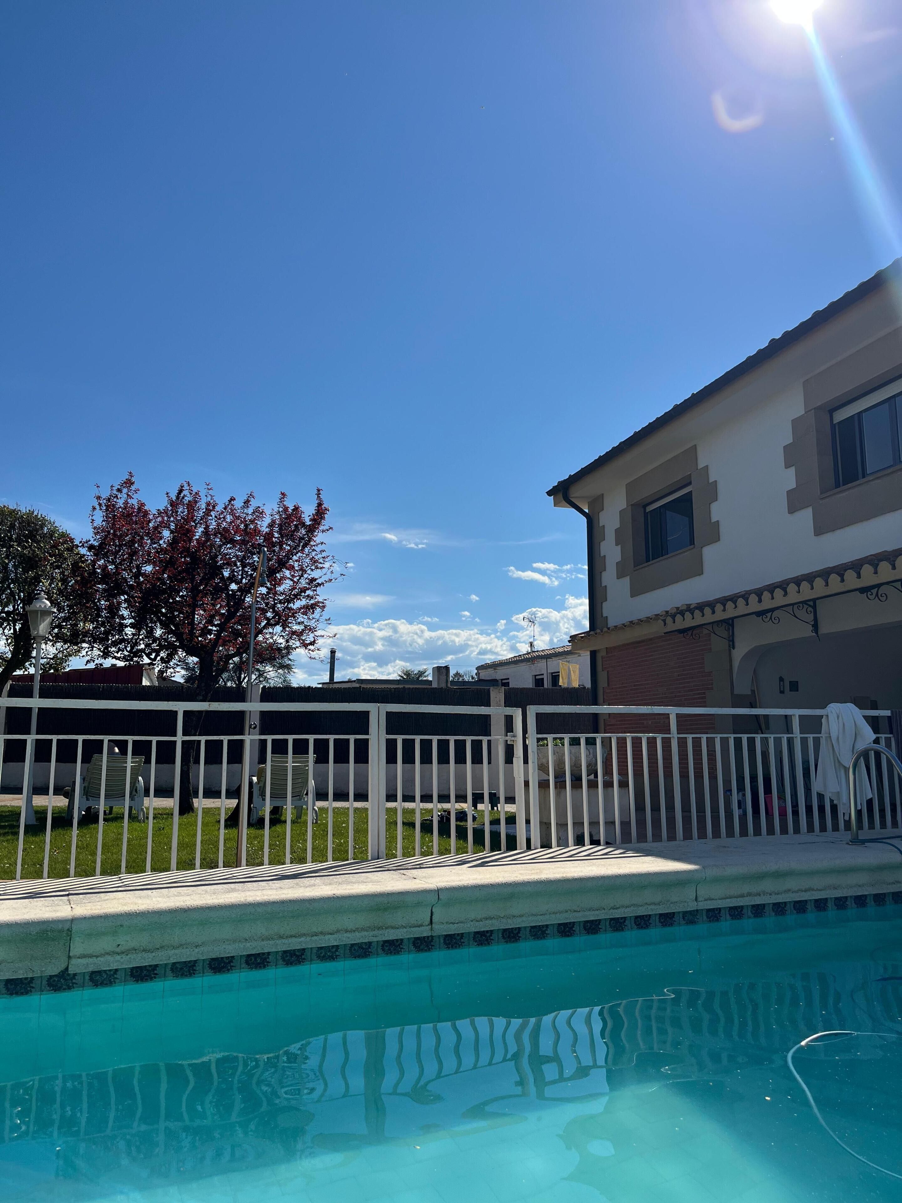 Una piscina con borde de azulejos, una valla blanca y un edificio con techo de tejas al fondo.