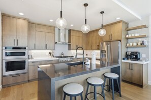 A modern kitchen with wooden cabinets, stainless steel appliances, a central island with stools, pendant lighting, and built-in shelves. - Sleeps 32 | 2 Hot Tubs + Sauna | Reservoir Views | Serenity Now + Pretty Good (Hideout)