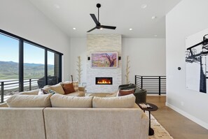 A modern living room with a fireplace, large windows, and a ceiling fan. - Sleeps 32 | 2 Hot Tubs + Sauna | Reservoir Views | Serenity Now + Pretty Good (Hideout)