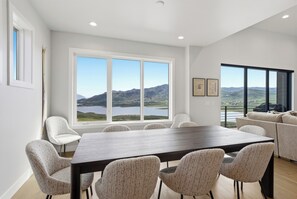 A modern dining area with a large table and chairs, large windows with a mountain view, and a sofa in the background. - Sleeps 32 | 2 Hot Tubs + Sauna | Reservoir Views | Serenity Now + Pretty Good (Hideout)