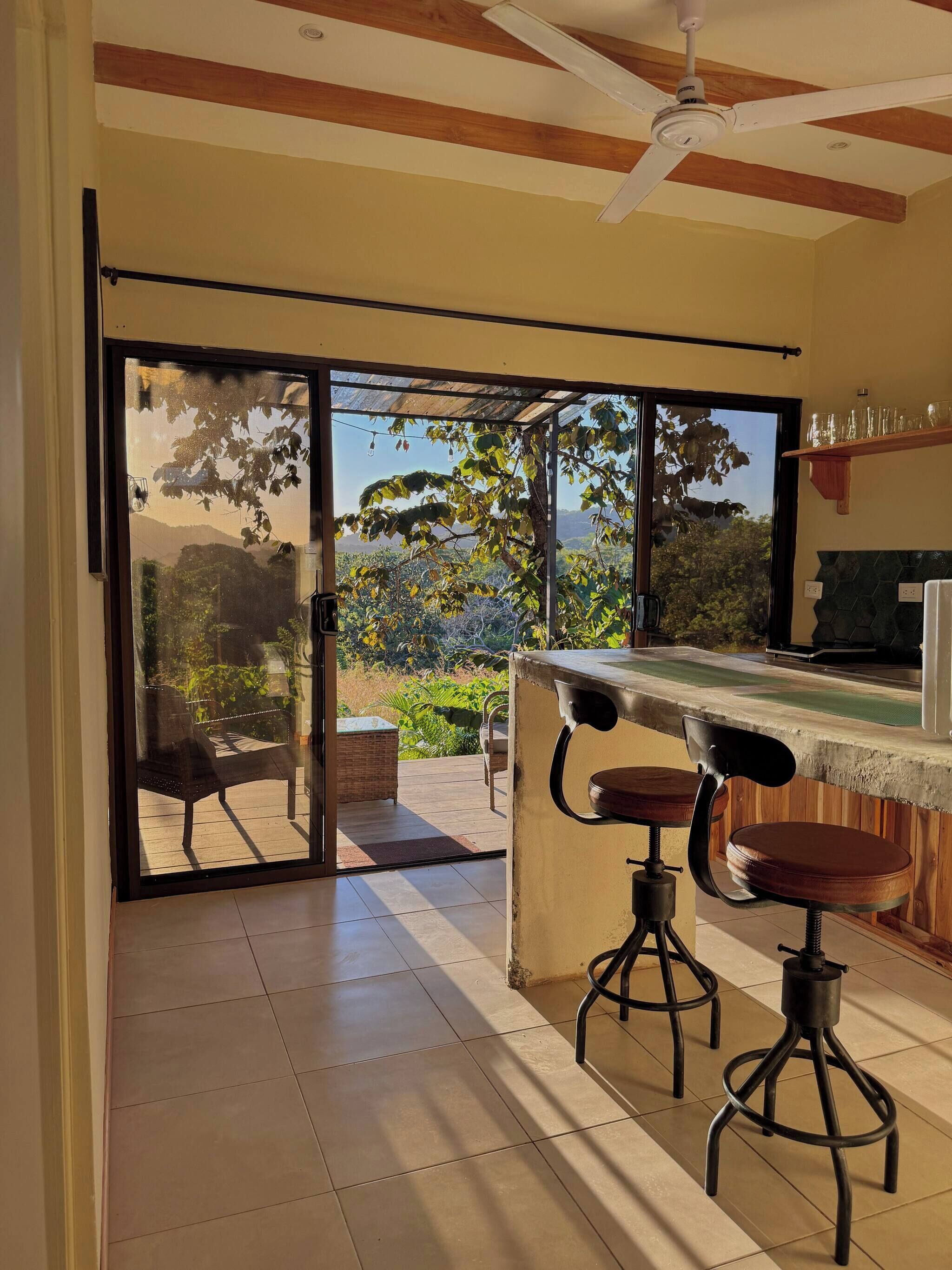 A room with a sliding glass door leading to an outdoor area, a ceiling fan, and a bar area with stools.