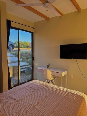 A bedroom with a bed, a desk, a chair, a television, and a sliding door leading to an outdoor area.