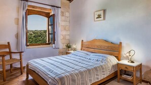 Ein Schlafzimmer mit einem hölzernen Bett, einem Nachttisch mit Lampe, einem Sessel, einem Fenster mit Vorhängen und einem gerahmten Bild an der Wand.