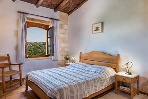 A bedroom with a wooden bed, a nightstand with a lamp, a chair, a window with curtains, and a framed picture on the wall. - Kamares – a restaured Venetian domain retreat (Macheri)